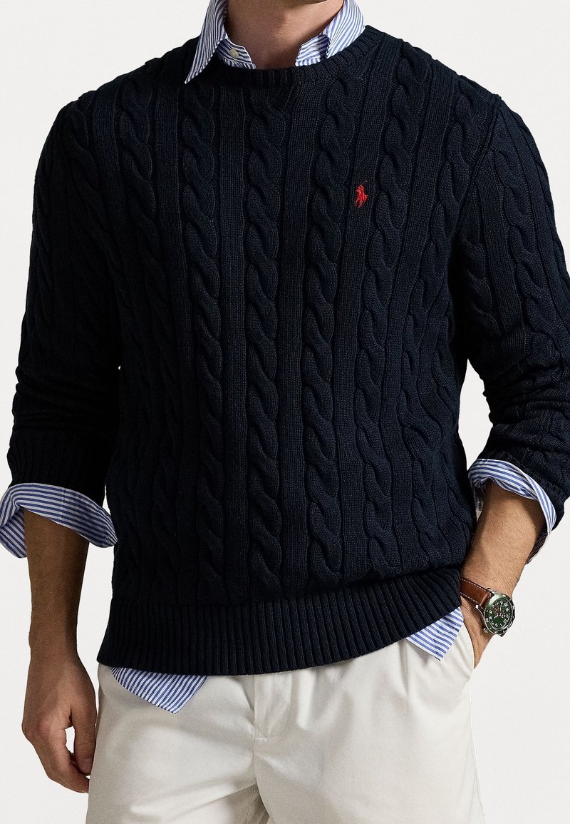 Homme portant un pull torsadé bleu marine avec un logo rouge sur une chemise à rayures bleues, un pantalon beige et une montre-bracelet marron avec un cadran vert.