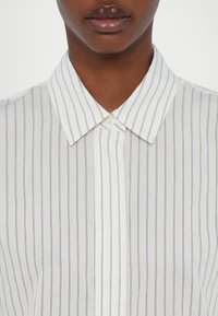 Bela srajca z velikimi navpičnimi modrimi črtami, z nazobčano ovratnikom in enim gumbom za zapiranje pri vratu. Gladek tekstil.