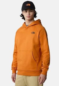 Sudadera naranja con un bolsillo tipo canguro, puños y dobladillo acanalados. Presenta un pequeño logo negro en el pecho. Se lleva con una gorra negra.