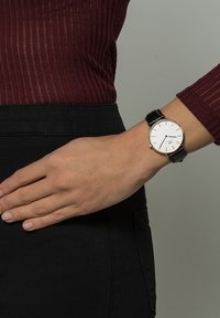 Kerek fehér óra minimalista dizájnnal, arany kiegészítőkkel és fekete bőrszíjjal, egy csuklón elhelyezve sötét blúz ellen.