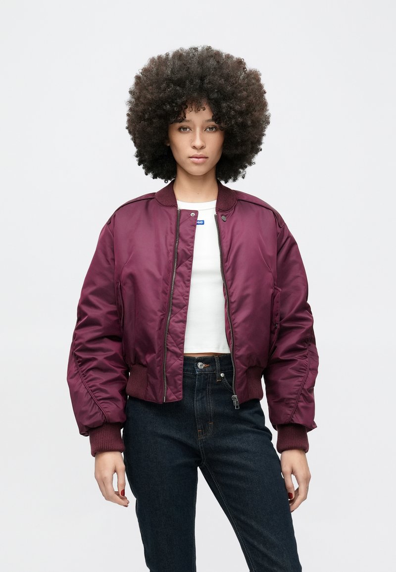 Bordos krāsas bumbvedēja jaka ar mirdzošu apdari, ribotām piedurknēm un rāvējslēdzēju priekšpusē. Kombinēta ar baltu topu un tumšām džinsa biksēm.