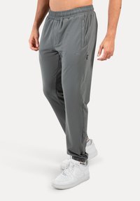 Grå træningsbukser med elastisk talje, sidelommer og lynlås-nederste del. Båret med hvide sneakers med sorte detaljer.