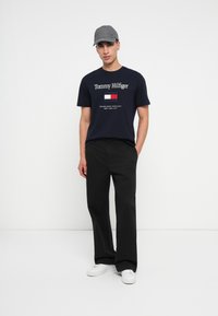 Νεαρός άνδρας φοράει γκρι καπέλο, μπλε T-shirt Tommy Hilfiger, μαύρο παντελόνι και λευκά αθλητικά παπούτσια, στέκεται σε έναν απλό λευκό φόντο.
