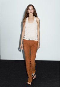 Beige linne med dekorerad fåll och byxor i brunt mocka. Modellen har på sig svarta flip-flops, med en minimalistisk bakgrund och naturlig hårtextur.