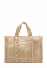 Beige tote bag with textured fabric, featuring plush edges, gold-tone studs, and embossed logo. Double handles for easy carrying.