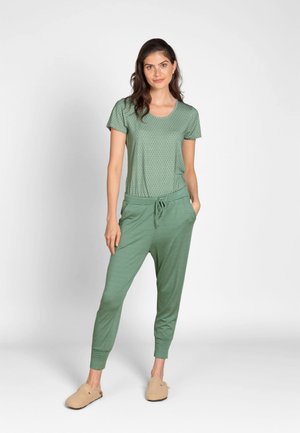 Woman wearing green patterned short-sleeve top and striped jogger pants with beige slippers, standing with one hand in pocket against white background.
