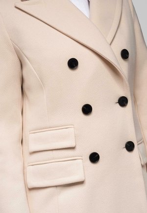 Beige dubbele-breasted jas met zwarte knopen, brede revers en twee klepzakken aan de linkerkant.