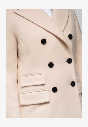 Abrigo de color beige cruzado con botones negros, amplios solapas y dos bolsillos de solapa en el lado izquierdo.