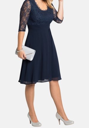Frau in einem knielangen marineblauen Spitzenkleid hält eine silberne Clutch, trägt silberne High Heels, ein Armband und eine Halskette vor hellem Hintergrund.