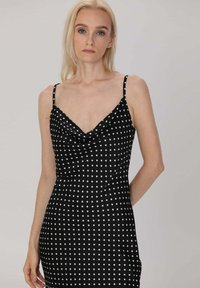 Robe noire à pois blancs avec un décolleté drapé, des bretelles fines, une silhouette ajustée et une texture de tissu lisse. Présente des pois blancs uniformément espacés.