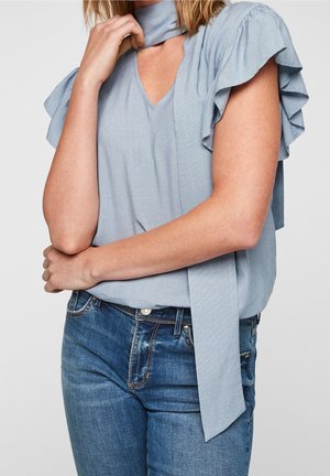 Femme portant un chemisier bleu clair à manches volantées avec un détail de lien au cou et un jean bleu, montrée de la moitié du torse au cou.