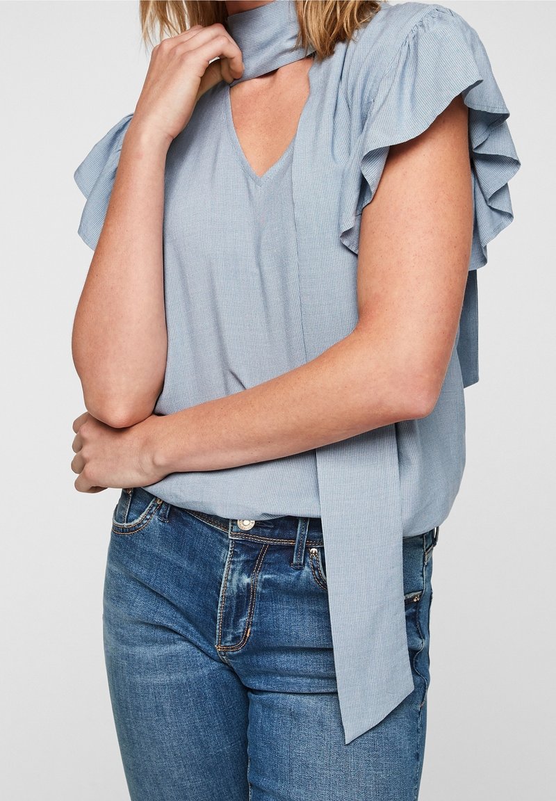 Femme portant un chemisier bleu clair à manches volantées avec un détail de lien au cou et un jean bleu, montrée de la moitié du torse au cou.