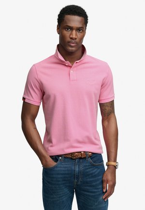 Uomo che indossa una polo rosa a maniche corte, jeans blu con cintura intrecciata e orologio, in piedi con una mano in tasca.