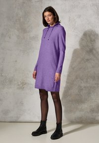 Abito lavorato a maglia viola con cappuccio e lacci, con maniche lunghe e vestibilità ampia, abbinato a stivali neri con lacci e collant neri.