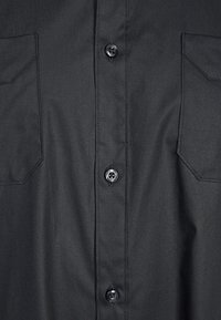 Camisa negra de botones con una textura suave, dos bolsillos en el pecho y botones en contraste en un diseño elegante y aerodinámico.