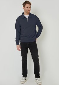 Marineblaue Zip-Up-Sweatjacke mit lockerer Passform, Kragen und gerippten Bündchen, kombiniert mit schwarzen Jeans und weißen Turnschuhen.