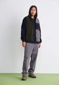 Kamizelka polarowa z oliwkowym zielonym panelem, szarymi i brązowymi akcentami, zamkiem błyskawicznym i małym logo. Noszona na czarnej bluzce z długim rękawem.