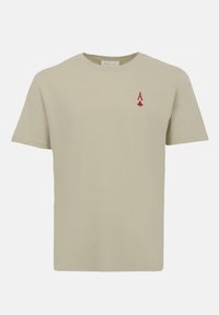 Μπεζ κοντομάνικη μπλούζα με ένα μικρό κόκκινο σύμβολο Άσσος Τριφύλλι από τράπουλα στο αριστερό στήθος.