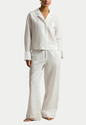 Kobieta ubrana w luźną, białą koszulę na guziki z kieszenią na piersi oraz pasujące szerokie spodnie, stojąca boso na jednolitym tle.