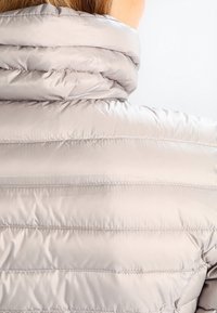 Jasnoszary puchowy kurtka z wysokim kołnierzem, pikowaną fakturą i poziomym przeszyciem, prezentująca gładkie, błyszczące wykończenie.