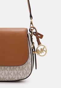 Beige and brown crossbody bag with "MK" logo pattern, leather flap, and gold circular MK charm hanging from strap attachment.