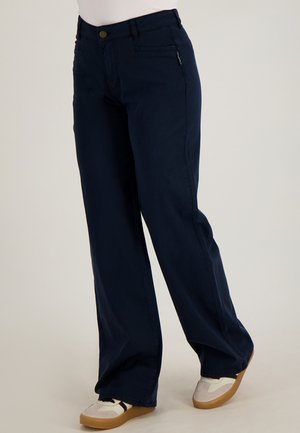 Persoon met losse donkerblauwe broek en witte sneakers met gumzolen, staande tegen een effen lichte achtergrond.