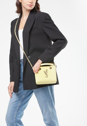 Donna che indossa un blazer nero e jeans azzurri chiari, tiene una piccola borsa gialla con tracolla a catena dorata e logo davanti.