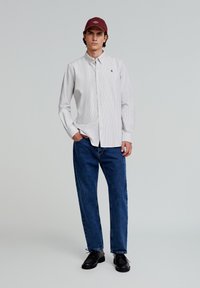Cămașă cu mânecă lungă, albă și gri cu dungi, blugi din denim albastru, pantofi negri, pălărie vișinie cu logo, model în picioare, fundal neutru.