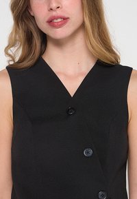 Vestito nero senza maniche con scollatura a V profonda, caratterizzato da tre bottoni e una silhouette aderente. Tessuto testurizzato con una finitura liscia.
