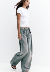 Mladá žena kráča, oblečená v priliehavom bielom tričku, nadmerných vyblednutých sivých džínsoch a čiernych žabkách, na bielom pozadí.