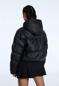 Veste noire matelassée avec capuche, présentant une finition lisse, un motif matelassé et un ourlet élastique. Portée sur une jupe noire.