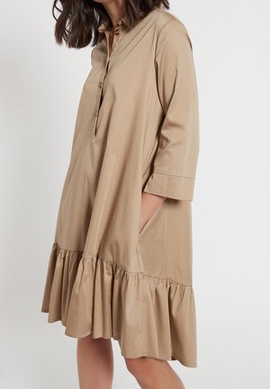 Mujer que lleva un vestido suelto de color marrón claro con mangas 3/4, cuello abotonado, bolsillos laterales y un dobladillo fruncido, de pie sobre un fondo blanco.