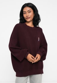 Burgundský oversized sveter s textúrovaným povrchom, spadnutými ramenami a rebrovanými manžetami. Na prednej strane sa nachádza malá nášivka loga.