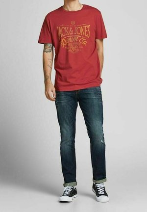 Punainen puuvillainen t-paita, jossa keltainen painettu logo "Jack & Jones Denim Co." Yhdistetty tummansinisiin farkkuihin ja mustiin lenkkareihin, joissa valkoiset pohjat.