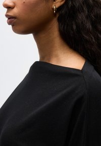 Προφίλ γυναίκας με μπούκλες, που φοράει μαύρη μπλούζα και μικρό χρυσό σκουλαρίκι σε απλό φόντο.