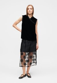 Svart ärmlös topp med volangkant vid halsringningen, kombinerad med en genomskinlig svart kjol med stora prickar. Modellen bär svarta ballerinaskor.