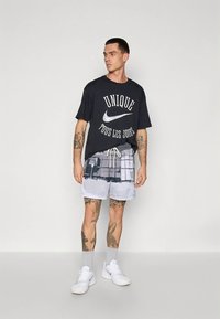 T-shirt preta de algodão com texto branco "UNIQUE TOUS LES JOURS" e logótipo da Nike; calções cinza claro com padrão geométrico; tênis brancos.