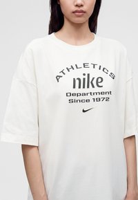 Γυναίκα φοράει μια λευκή φαρδιά μπλούζα Nike με μαύρο κείμενο "Athletics nike Department Since 1972" και μικρό λογότυπο Nike swoosh.