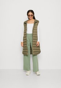 Gilet à capuche en olive vert foncé, avec un design matelassé horizontal. Assocé à un pantalon large vert clair et des baskets blanches.