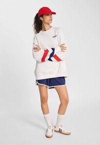 Vitt sweatshirt med röda och marinblå diagonala ränder, marinblå shorts, röd keps och vita strumpor. Sneakers har bruna sulor och diskret märkning.