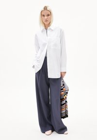 Weißes Button-Up-Hemd mit einer Brusttasche, kombiniert mit lässig geschnittenen dunkelblauen Hosen. Ein mehrfarbig gestreifter Strickschal ist über den Arm drapiert.