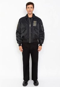 Czarna kurtka bomberka o satynowym wykończeniu, zapinana na zamek, z dwoma przednimi kieszeniami. Posiada ozdobną naszywkę na piersi oraz ściągacze na mankietach i u dołu.