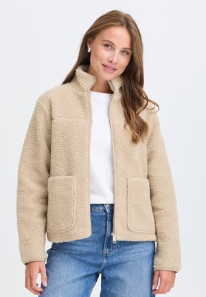 Femme portant une veste polaire beige par-dessus une chemise blanche et un jean bleu, souriante avec des cheveux ondulés lâchés sur un fond uni.