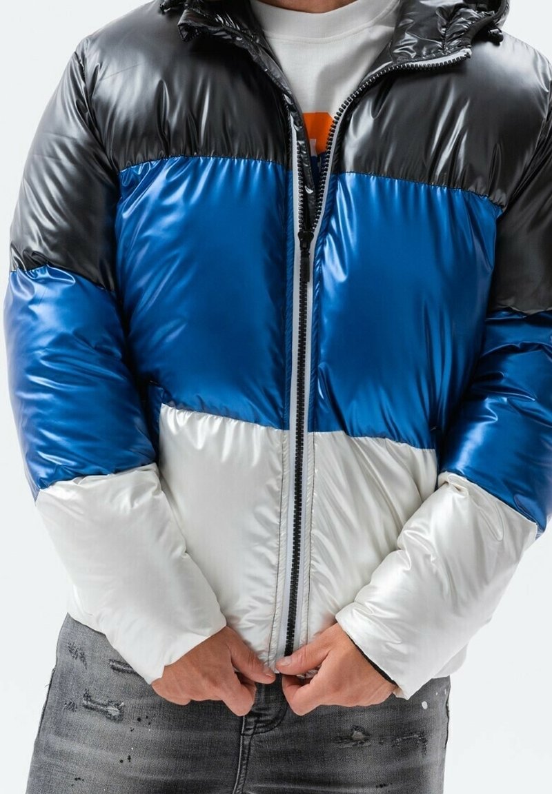 Gepolsterte Jacke mit glänzendem Finish, die schwarze, blaue und weiße Farbblöcke aufweist. Reißverschlussverschluss und Kapuzen-Design. Darunter getragene Jeans.