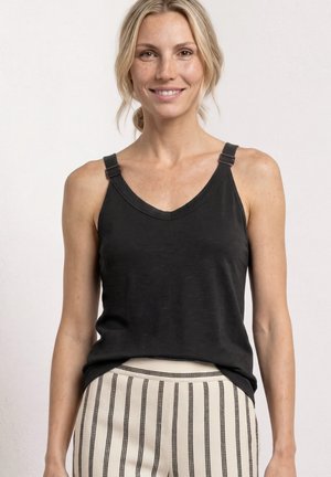 Femme souriante aux cheveux blonds, portant un haut noir sans manches et un pantalon crème à rayures verticales noires, debout devant un fond clair uni.