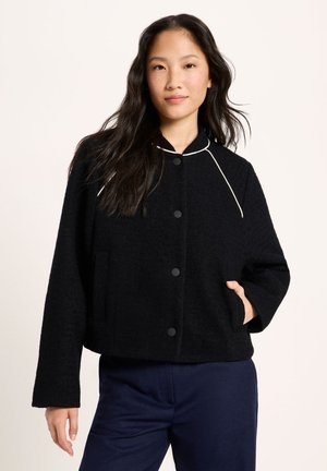 Woman with long dark hair wearing a black buttoned jacket with white piping and navy blue pants, hand in jacket pocket, neutral background.