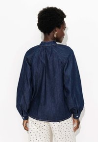 Chemise en denim bleu foncé, avec des manches ballons, un col froncé et une taille cintrée, associée à un pantalon blanc à motifs.