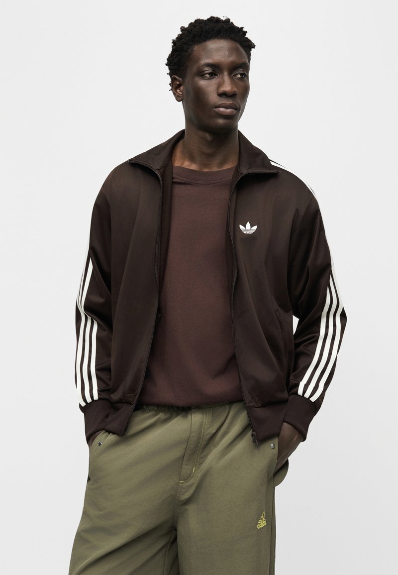 Modello maschile che indossa una giacca sportiva Adidas marrone scuro con strisce bianche e logo, maglietta marrone e pantaloni Adidas verdi oliva con logo.
