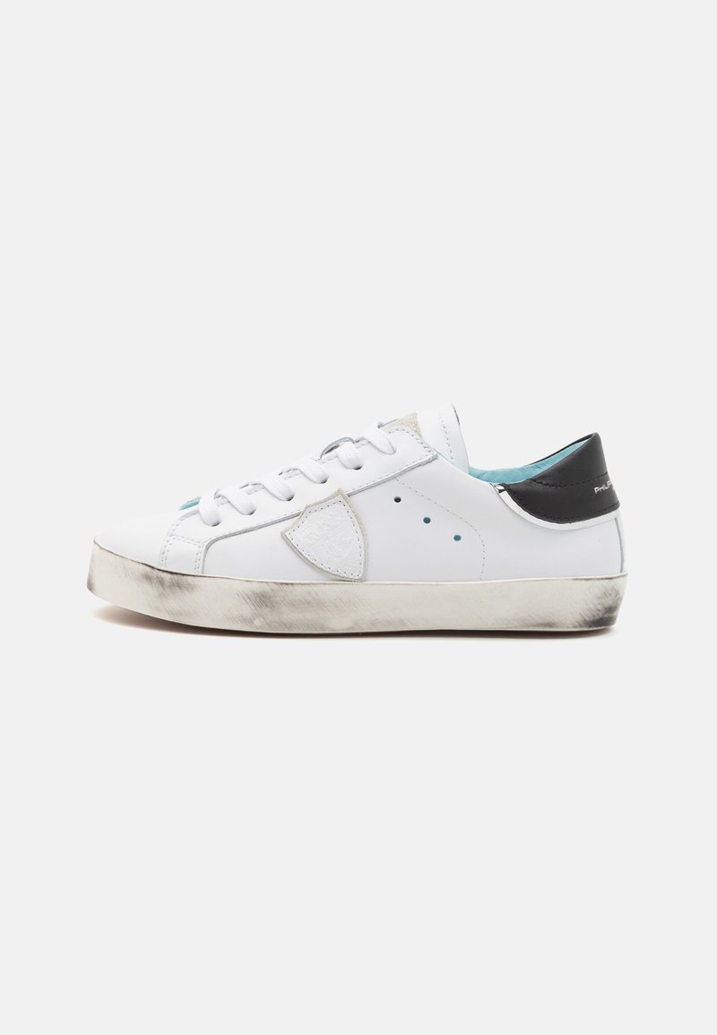 White leather sneaker with a black heel accent, perforated details, round toe, and textured sole showing wear, featuring blue interior lining.