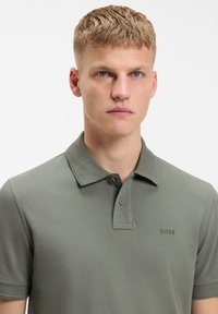 Jeune homme aux cheveux blonds courts portant un polo BOSS vert olive, face à l'avant avec une expression neutre, fond blanc uni.